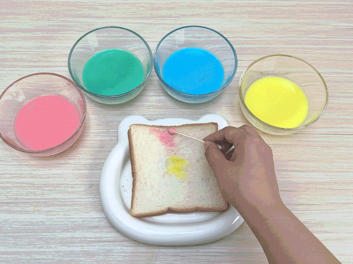 A slice of white bread rests on a small, white, bear-shaped plate on a wooden table. Four small glass bowls, each containing a different colored liquid – pink, green, blue, and yellow – are positioned above the bread. A hand uses cotton swabs to apply the colored liquids onto the bread, creating a colorful design. This video likely demonstrates a creative food decorating activity, possibly for children.