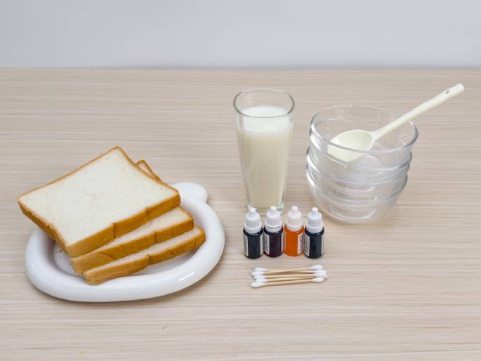 Ingredients and tools for a food-related activity are arranged on a light wood-grained table. Three slices of white bread are stacked on a white, bear-shaped dish. Next to the bread are a glass of milk, a stack of small clear glass bowls containing a white substance (possibly sugar or powdered milk) with a white spoon resting on top, four small bottles of food coloring in various colors (black, red, orange, and dark blue), and a group of cotton swabs. The setup suggests a project involving decorating or coloring the bread, possibly using the milk and food coloring as paint.