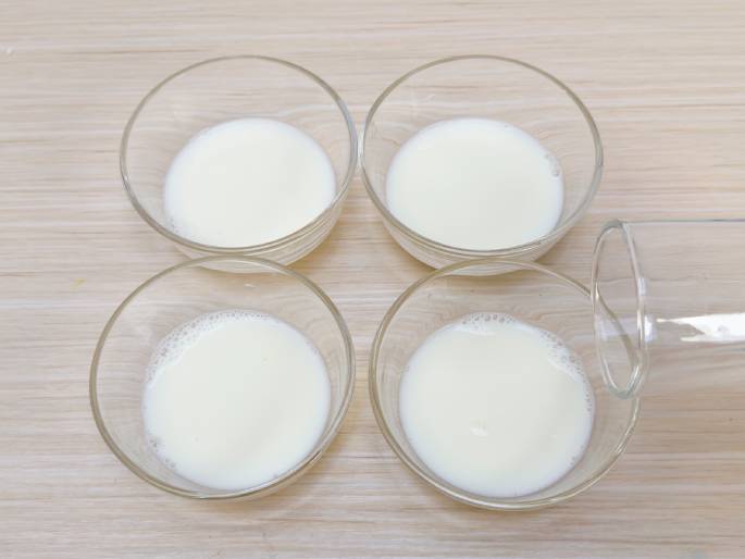 Four small, clear glass bowls each containing milk are arranged on a light wood-grained surface. A drinking glass, likely the source of the milk, lies on its side, partially overlapping the bottom right bowl. The milk in the bowls appears freshly poured, with a few small bubbles visible in the lower two bowls. This image likely depicts a preparation step for a recipe or activity involving milk.