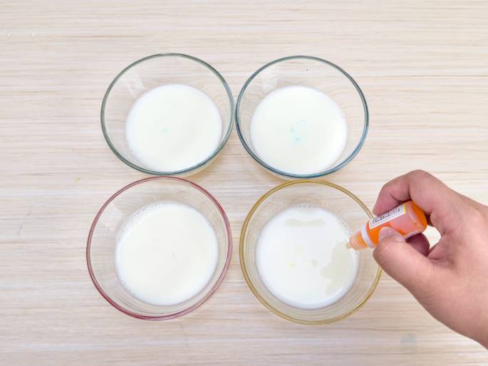 A hand adds orange food coloring from a small bottle to a bowl of milk. Four clear glass bowls, each containing milk, are arranged on a light wood-grained surface. A small drop of blue coloring is visible in the top right bowl, and a few drops of orange coloring are dispersing into the bottom right bowl. The other two bowls of milk remain uncolored. This image likely demonstrates the beginning stages of a food coloring experiment or a recipe involving colored milk.