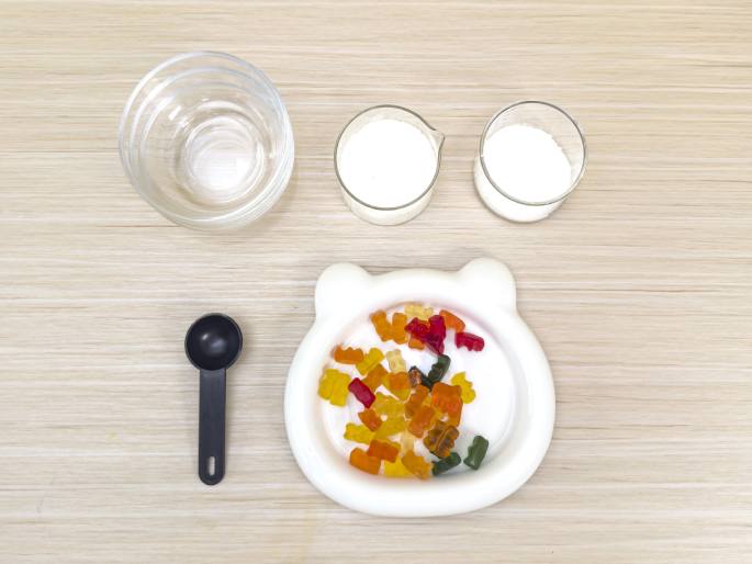 Ingredients and tools are laid out on a light wooden table, seemingly for a gummy bear-related activity. A white bear-shaped dish holds a small pile of colorful gummy bears. Above the dish are an empty clear glass, presumably for water, and two smaller clear containers filled with a white substance, likely sugar or a similar ingredient. A small black measuring spoon lies to the left of the bear dish. The arrangement suggests a candy-making or science experiment involving gummy bears.