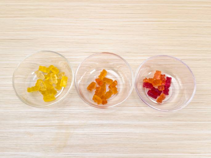Three clear glass bowls are arranged on a light wood-grained surface. Each bowl contains a small handful of gummy bears. The gummy bears are grouped by color, with yellow bears in the left bowl, orange bears in the middle bowl, and red bears in the right bowl. This image likely depicts a sorting activity or the ingredients for a recipe or experiment involving gummy bears.