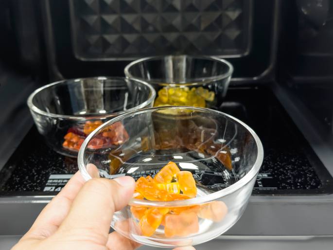 A hand holds a clear glass bowl of orange gummy bears in the foreground, just inside the open door of a microwave oven. Two other glass bowls, containing yellow and red gummy bears, are visible further inside the microwave on the rotating glass plate. The microwave interior is black with a textured pattern. This image likely depicts a moment in an experiment or activity involving heating gummy bears in a microwave.