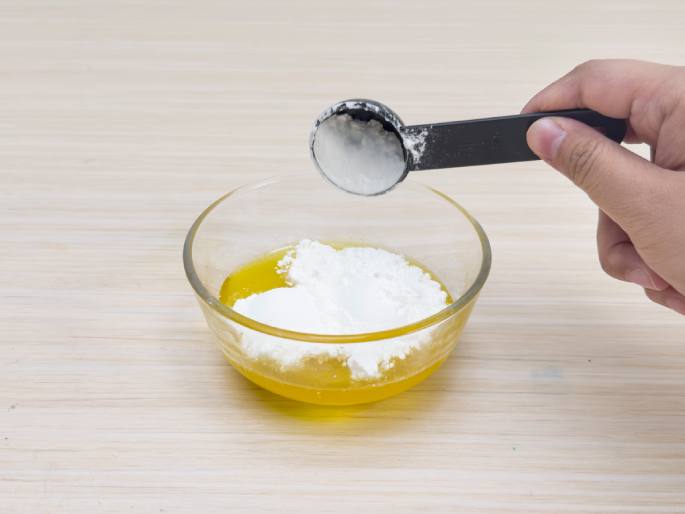 A hand holds a black measuring spoon filled with white powder, poised above a clear glass bowl. The bowl contains a yellow liquid and some of the white powder has already been added. The bowl sits on a light wood-grained surface. This image likely depicts someone adding a powdered ingredient, possibly cornstarch or flour, to a liquid, possibly oil or melted butter, as part of a recipe.