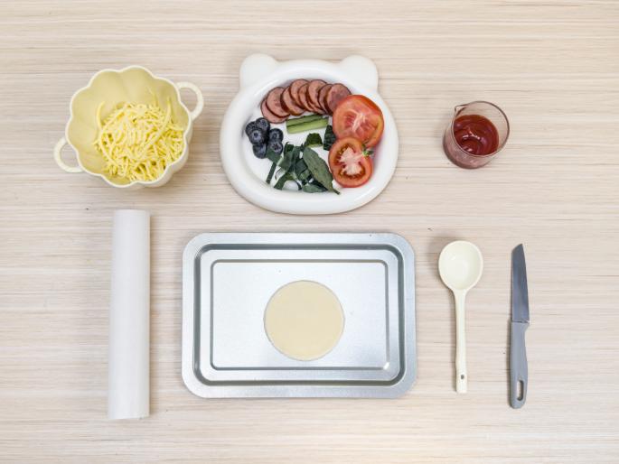 An overhead flatlay image shows ingredients and tools for making mini pizzas arranged on a light wood-grained surface. A small, off-white bowl contains shredded cheese. A white bear-shaped dish holds sliced pepperoni, tomato slices, blueberries, and spinach leaves. A small, clear container holds tomato sauce. A roll of parchment paper lies near a small baking sheet with a single small pizza crust on it. A white spoon and a gray butter knife complete the scene. This image appears to be set up for a child or for demonstrating a simple pizza-making activity.