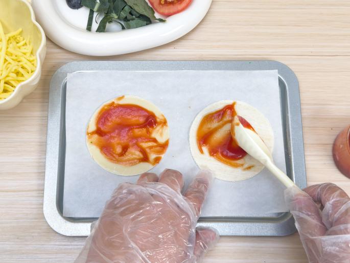 Two hands wearing clear plastic gloves are preparing small pizza crusts on a baking sheet lined with parchment paper. One hand holds a small white spatula and is spreading tomato sauce onto a crust. The other hand steadies the baking sheet. A second crust, already spread with sauce, sits next to it. A bowl of shredded cheese and other ingredients are visible in the background on a light wooden table, suggesting this is part of a pizza-making process.