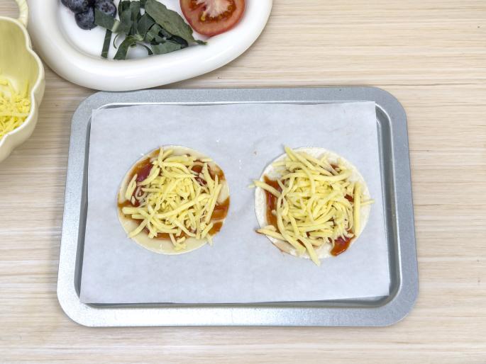 Two small pizza crusts topped with tomato sauce and shredded cheese sit on a baking sheet lined with parchment paper. The baking sheet rests on a light wood-grained table. Other pizza toppings, such as sliced tomatoes, blueberries, and spinach, are visible in a dish in the background, indicating that these pizzas are in the process of being assembled.