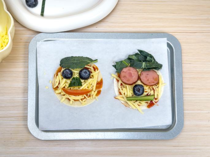 Two small pizzas decorated as faces are placed on a baking sheet covered with parchment paper, which sits on a light wooden table. Both pizzas have tomato sauce and shredded cheese. The first pizza uses spinach for hair, blueberries for eyes, a cucumber slice for a nose, and shredded tomato for a mouth. The second pizza uses spinach for hair, pepperoni slices for eyes, a blueberry for a nose, and a cucumber stick for a mouth. These fun pizzas are likely designed for children.