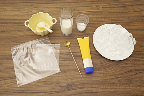 Several items are arranged on a dark brown wooden surface, clearly laid out for a recipe. These include two clear plastic bags of differing sizes, a small pale yellow bowl with a spoon, a glass containing milk, a small cup with what appears to be salt, a small gold spoon, a tube of yellow substance (possibly ice cream base or cream), and a plate piled with ice cubes. The overall impression is of preparation for a homemade ice cream recipe, possibly involving a no-churn method given the absence of an ice cream maker. The scene is well-lit and neatly presented.
