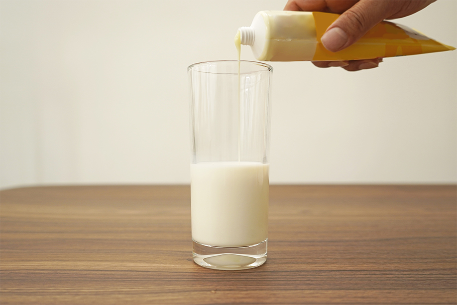A hand is squeezing a tube of pale yellow cream into a tall, clear glass containing milk. The cream is pouring smoothly into the milk, creating a creamy mixture. The glass sits on a light brown wooden surface. The background is a softly lit, out-of-focus off-white wall. The image clearly shows a step in a recipe, likely for homemade ice cream, focusing on the addition of a key ingredient to the milk base.