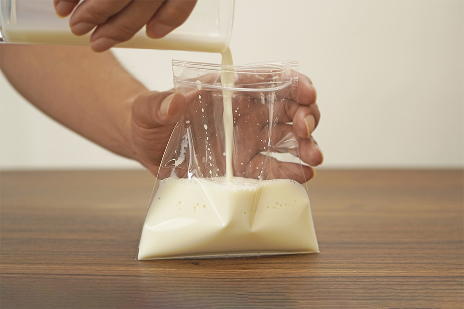A hand carefully pours milk from a glass into a clear plastic freezer bag held open by another hand. The milk flows steadily into the bag, which is positioned on a dark brown wooden surface. The background is a softly lit, out-of-focus off-white wall. The image clearly shows a step in a no-churn ice cream recipe, focusing on the careful transfer of milk into a freezer bag for subsequent freezing and mixing.