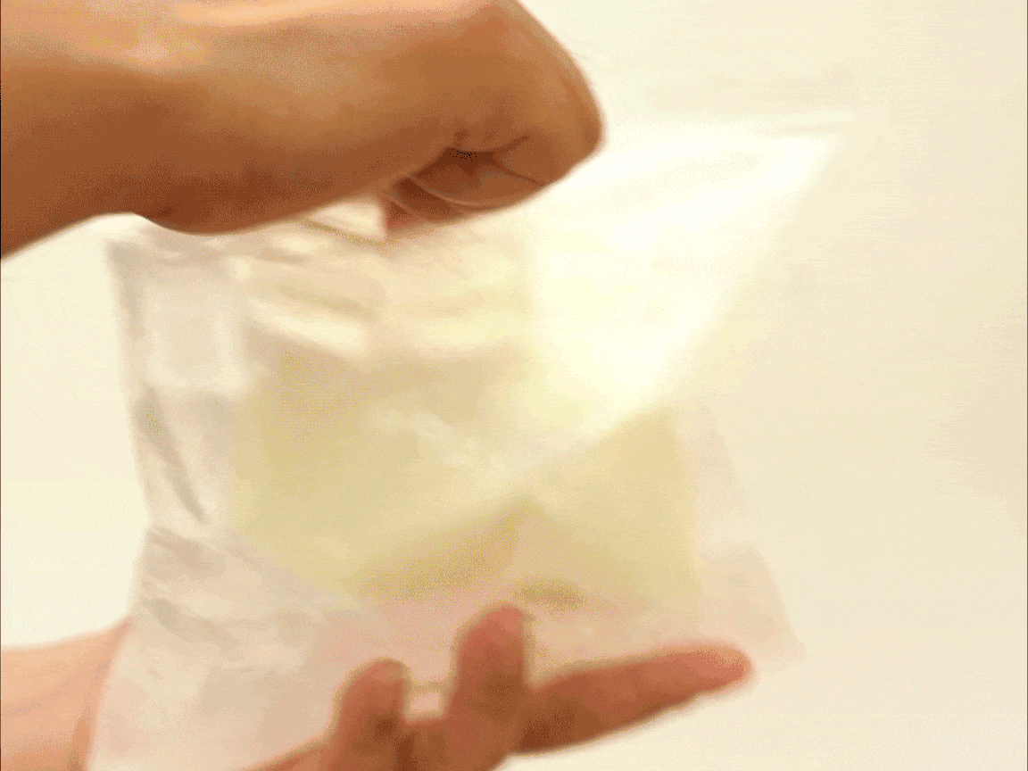 A hand is shown gently manipulating a clear plastic freezer bag containing a pale yellow, creamy mixture, likely the ice cream base. The bag is being squeezed and manipulated, suggesting the process of mixing the ingredients within. The background is a softly lit, out-of-focus off-white wall. The image focuses on the action of mixing the ice cream base within the bag, a key step in a no-churn ice cream recipe. The overall impression is one of a simple, hands-on method.