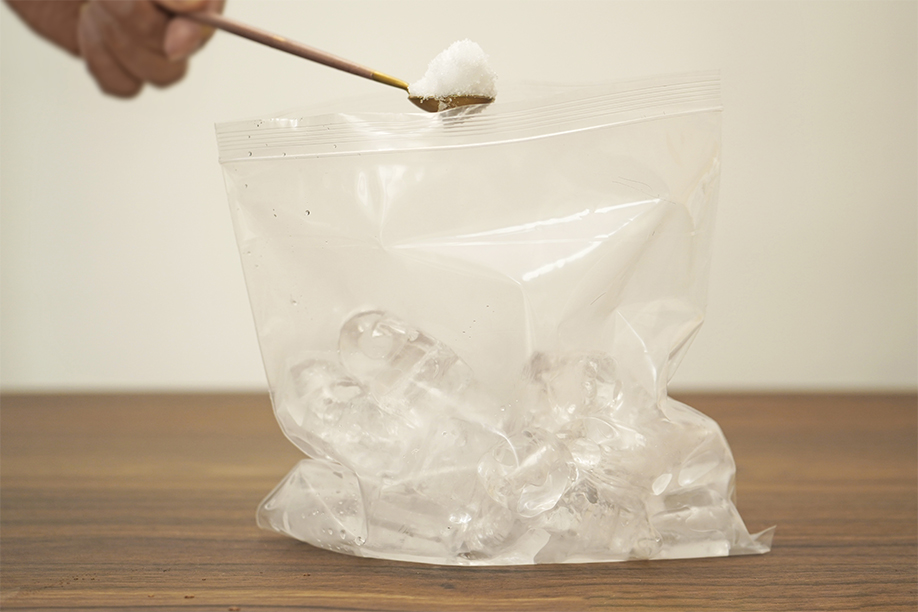 A hand uses a gold spoon to add a spoonful of salt to a clear plastic freezer bag already containing ice cubes. The bag rests on a dark brown wooden surface. The background is a softly lit, out-of-focus off-white wall. The image clearly shows one step in a no-churn ice cream recipe, highlighting the addition of salt to the ice bath which will help to freeze the ice cream mixture more quickly.