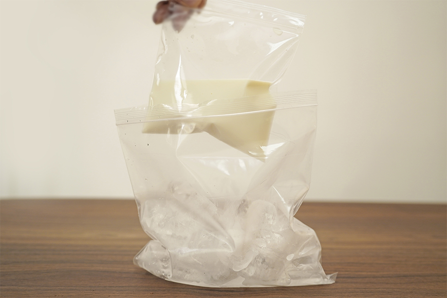 A hand carefully places a smaller, clear plastic bag containing a creamy, off-white mixture into a larger, clear plastic bag filled with ice cubes. The smaller bag is positioned on top of the ice, suggesting a method for chilling or freezing the mixture. The larger bag sits on a dark brown wooden surface. The background is a softly lit, out-of-focus off-white wall. The image depicts a step in a no-churn ice cream recipe, emphasising the process of chilling the ice cream base within an ice bath for rapid cooling.