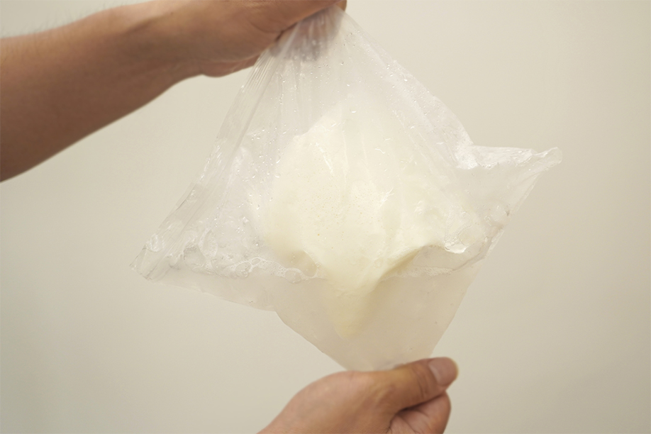 Two hands hold a clear plastic freezer bag containing a creamy, off-white mixture, possibly ice cream base. The bag is slightly puffed up, suggesting the mixture has expanded slightly. The background is a plain, off-white wall, drawing focus to the bag and its contents. The image shows a stage in a no-churn ice cream recipe, where the mixture has been chilled and is ready for the next step in the process. The overall impression is one of a simple, homemade method.