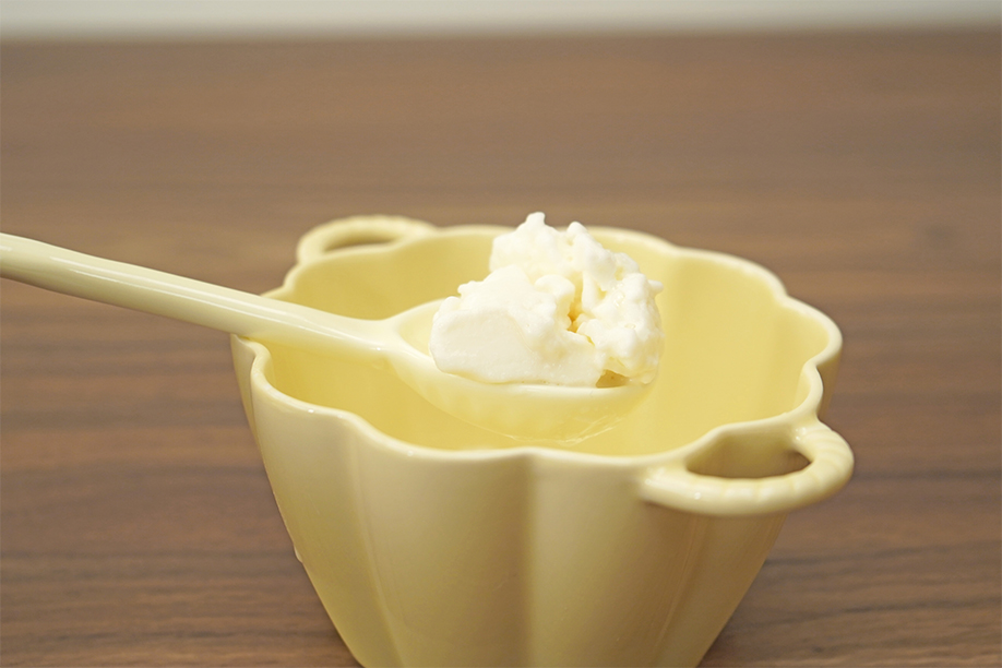 A pale yellow bowl sits on a wooden surface, partially filled with what appears to be the remnants of a no-churn ice cream mixture. A pale yellow spoon holds a small portion of the now-frozen ice cream, showcasing its creamy texture. The focus is on the smooth, white ice cream, highlighting the successful outcome of the homemade recipe. The overall setting is clean and simple, emphasising the delicious result of the process.