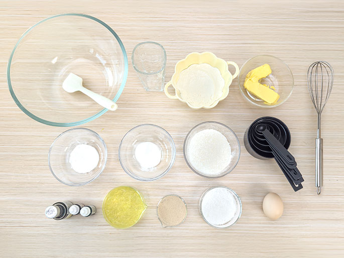 An overhead shot displays an array of baking ingredients and tools arranged on a light wood-grain surface. The items include a large clear glass mixing bowl with a white spatula, a clear glass, a small pale yellow bowl containing flour, a small clear glass bowl with butter, a metal whisk, two small clear glass bowls with white powders (likely baking soda and baking powder), a larger clear glass bowl with flour, a set of black measuring cups and spoons, a small clear measuring cup or beaker with a yellow liquid (possibly oil), a small clear measuring cup with brown sugar, a small clear bowl with a white powder (possibly sugar), and a single egg. The arrangement suggests preparation for baking.