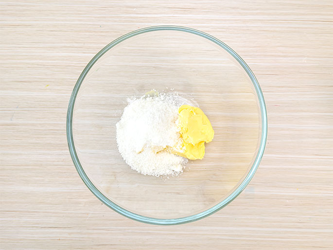 A clear glass mixing bowl sits on a light wood-grain surface. Inside the bowl, a small mound of white granulated sugar sits next to a pat of yellow butter. The ingredients appear ready to be creamed together, likely as a first step in a baking recipe.