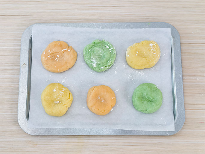 Six unbaked cookies, in three different colors, are arranged on a baking sheet lined with parchment paper. The colors are pale yellow, light orange, and light green, with two cookies of each color. They are evenly spaced and lightly dusted with powdered sugar. The image shows the cookies prepared and ready to be baked.