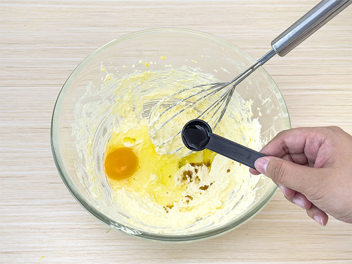 A clear glass bowl, containing a creamed mixture of butter and sugar, sits on a light wooden surface. A whole egg and a small amount of vanilla extract, held in a black measuring spoon by a hand, are being added to the mixture. A whisk is also in the bowl, implying the ingredients are about to be combined.