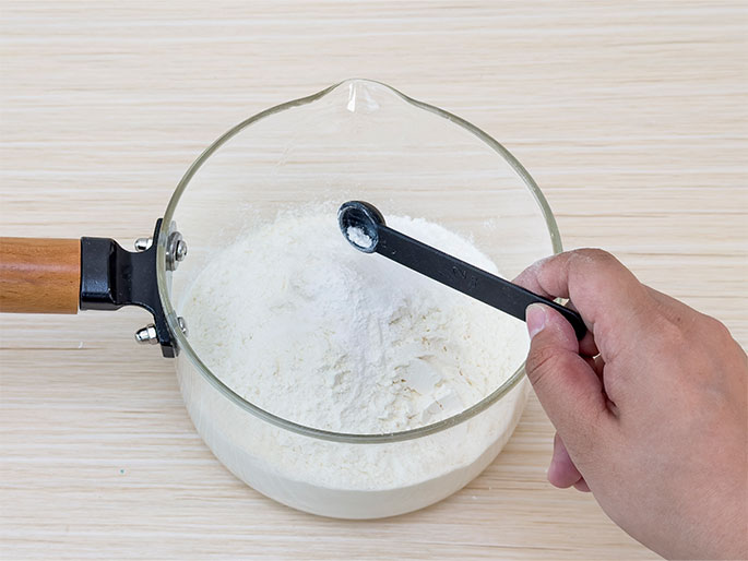A clear glass bowl with a wooden and black handle, resembling a small saucepan, sits on a light wood-grain surface. Inside the bowl is flour, and a hand holds a black measuring spoon containing a small amount of white powder, likely baking powder or baking soda, over the flour. This suggests the dry ingredients are being prepared for a baking recipe.