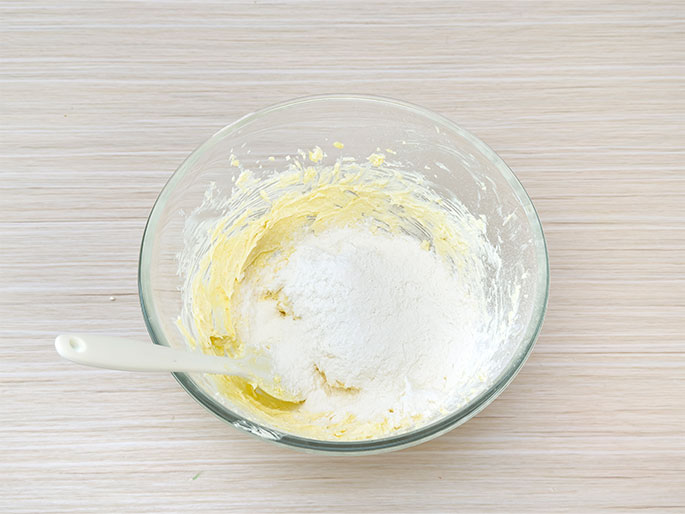 A clear glass mixing bowl sits on a light wood-grain surface. Inside, a pale yellow creamed mixture, likely butter and sugar, has a mound of white flour on top. A white spatula rests in the bowl, indicating the flour is about to be incorporated into the wet ingredients. This image depicts a stage in preparing a batter or dough.