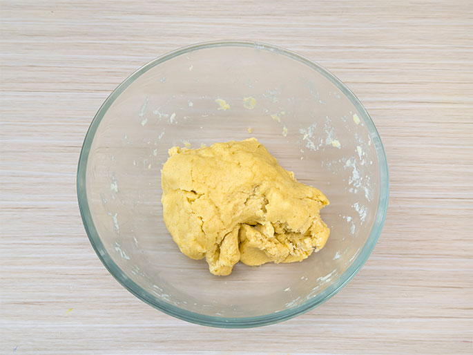 A clear glass mixing bowl rests on a light-colored, wood-grained surface. Inside the bowl is a mass of pale yellow dough, gathered together but still showing some folds and creases, indicating it has recently been mixed and is ready for the next stage of preparation, such as chilling or rolling.