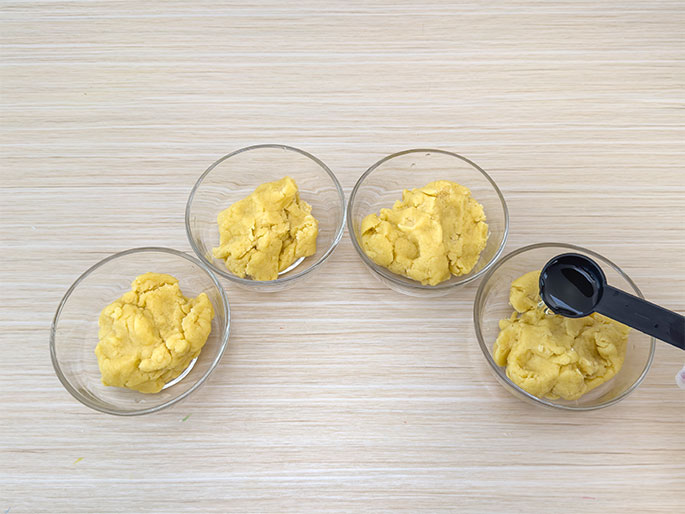 Four clear glass bowls, each containing a portion of pale yellow dough, are arranged in a semi-circle on a light wood-grain surface. A black measuring spoon, filled with a dark liquid, likely a food coloring or flavoring extract, is being poured into one of the bowls of dough. This image suggests the dough is being divided and prepared for individual flavoring or coloring.