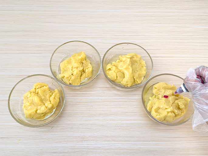Four clear glass bowls are arranged in a semi-circle on a light wood-grain surface. Each bowl holds a portion of pale yellow dough. A hand wearing a clear plastic glove is adding drops of red food coloring to one of the dough portions. The other three portions remain uncolored, indicating they will likely be colored differently or left plain. This image shows the process of preparing dough for colorful baked goods.