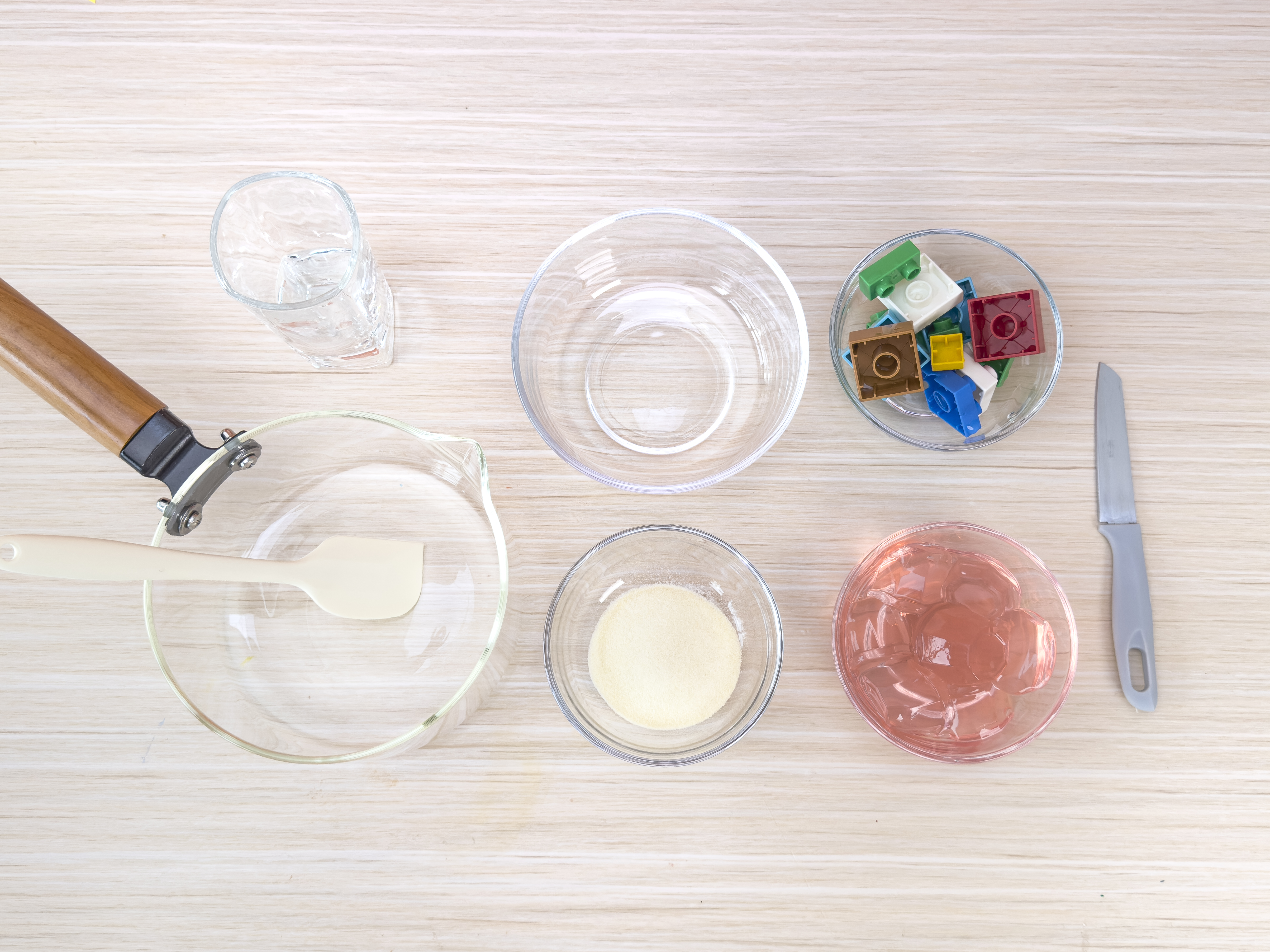 An overhead shot displays the ingredients and tools for making LEGO brick-shaped jellies. Arranged on a light wood-grain surface are various clear glass bowls and a glass. One bowl contains a pile of powdered gelatin, another holds a small amount of pre-made pink jelly, and a third has a collection of colorful LEGO bricks. A glass filled with water, a small glass pot with a wooden handle and a white spatula inside, and a silver butter knife complete the scene. This flat lay clearly presents the elements needed for this recipe.