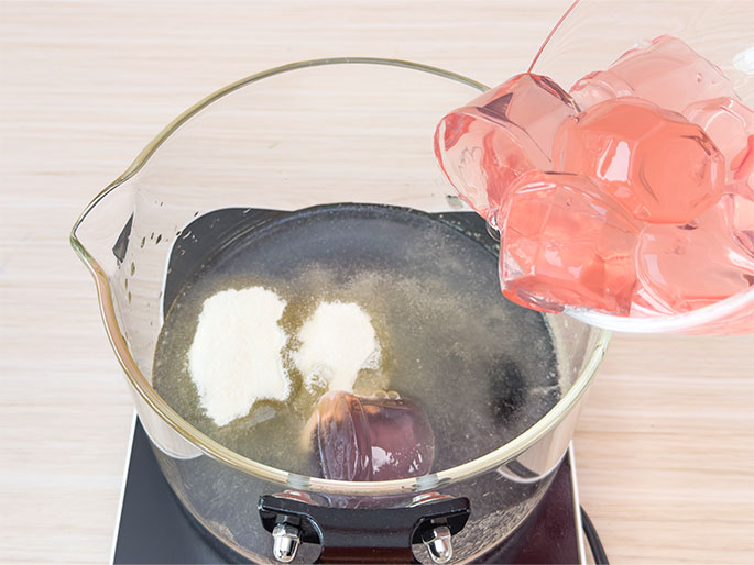 Pink jelly cubes are being poured from a clear container into a glass pot containing water and powdered gelatin. The pot sits on a black, electric burner. The process of dissolving gelatin and combining it with pre-made jelly is depicted here, likely a step in a recipe for creating layered or molded jellies.