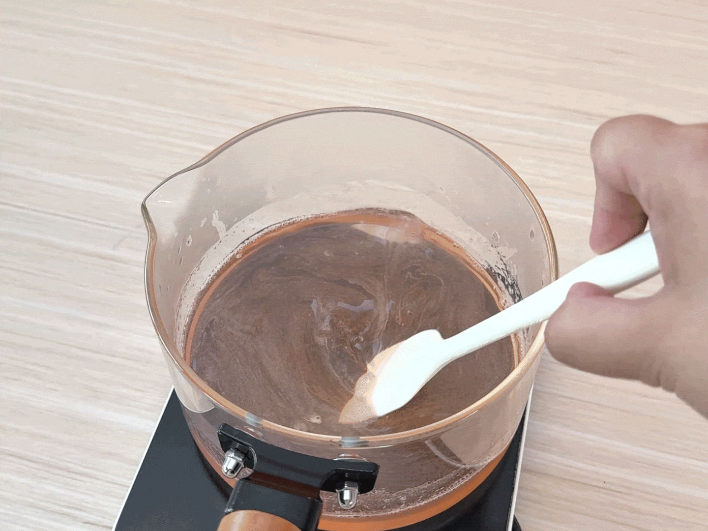 A hand uses a white spatula to stir a dark, brownish-red liquid in a clear glass pot on a black electric burner. The liquid appears to be a mixture of ingredients, possibly melting or dissolving, as it's being heated and stirred. The image suggests a cooking or candy-making process.