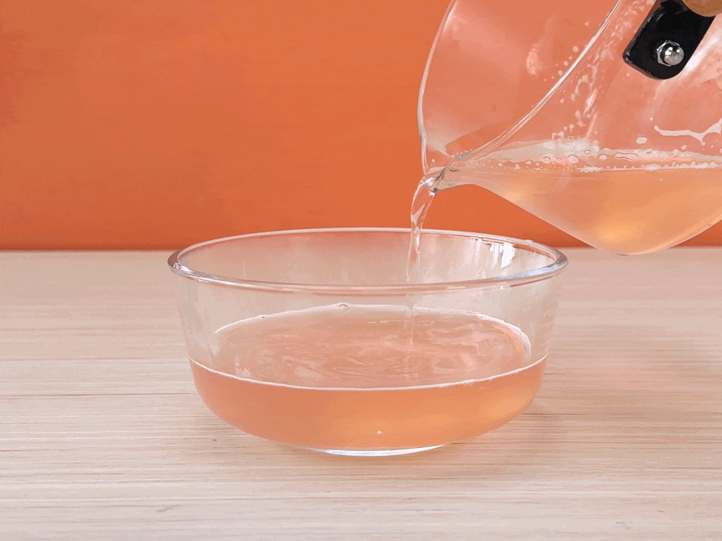 Against a vibrant orange backdrop, a light pinkish-orange liquid is being poured from a clear glass pot with a handle into a clear glass bowl. The liquid flows smoothly, and the bowl already contains a small amount of the same liquid. The scene likely depicts a step in a food preparation process, perhaps involving jelly or a similar dessert. The light wood-grain surface below the bowl adds a natural element to the composition.