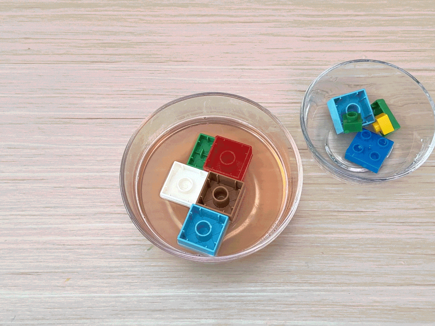 On a light wooden surface, two clear glass bowls are visible. One bowl is partially filled with a light orange liquid, and several small, colorful LEGO bricks are submerged in it. A second bowl sits next to it, containing a few more LEGO bricks, but without any liquid. This image likely illustrates a step in making LEGO-shaped jellies, where the molds are being prepared.