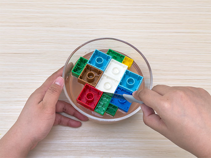 Two hands hold a clear glass bowl filled with a light brown liquid on a light wood-grain table. Colorful LEGO bricks are partially submerged in the liquid, arranged in a grid-like pattern. One hand uses a small, grey spatula to carefully adjust the position of a white LEGO brick within the liquid. This image suggests a crafting or culinary activity, possibly involving embedding the LEGOs in a gelatin or resin mixture.