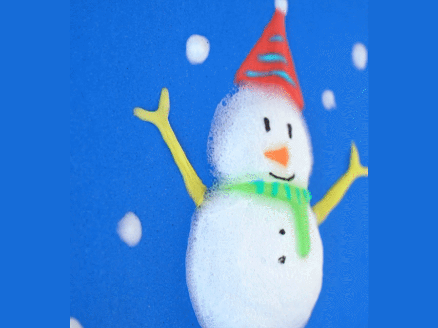 A foamy snowman is depicted on a blue background, adorned with a red and blue striped party hat and a light green scarf. Small, white foam dots around the snowman represent snowflakes. A finger pokes the snowman's midsection, creating a small indentation in the foam.
