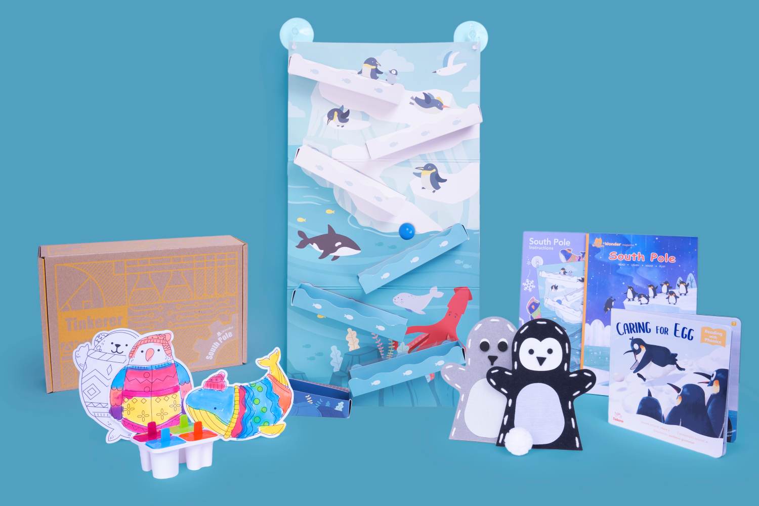 A collection of South Pole-themed educational toys and materials is displayed against a teal background. The central item is a vertical game board depicting penguins on ice floes above an ocean scene with a killer whale, squid, and other marine life. A light blue ball is visible mid-descent on the game's cascading chutes. In the foreground, colorfully decorated cutouts of a penguin, polar bear, and whale are attached to a white popsicle mold base. To the right, two felt penguin puppets and a white pom-pom snowball are shown alongside two small booklets titled South Pole Instructions, South Pole - Learn, Play, Make, and Caring for Egg - Reading & Phonics. A cardboard box labeled Tinkerer - South Pole is visible to the left. This arrangement suggests a comprehensive learning kit designed for children.