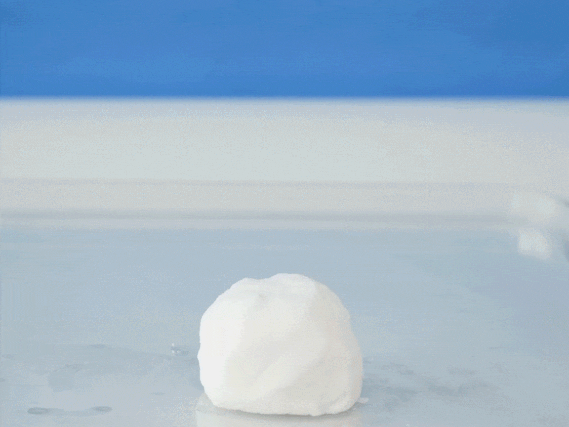 A small snowman is constructed from three white balls of material, likely dough or modeling clay, against a blue backdrop. Brown cardboard arms, two pink candy heart buttons, and a small red conical hat are added. A fuzzy orange scarf and googly eyes complete the snowman's festive look. Finally, a clear liquid, possibly water, is poured over the snowman, causing it to dissolve into a foamy white substance that fills the clear container it sits in.