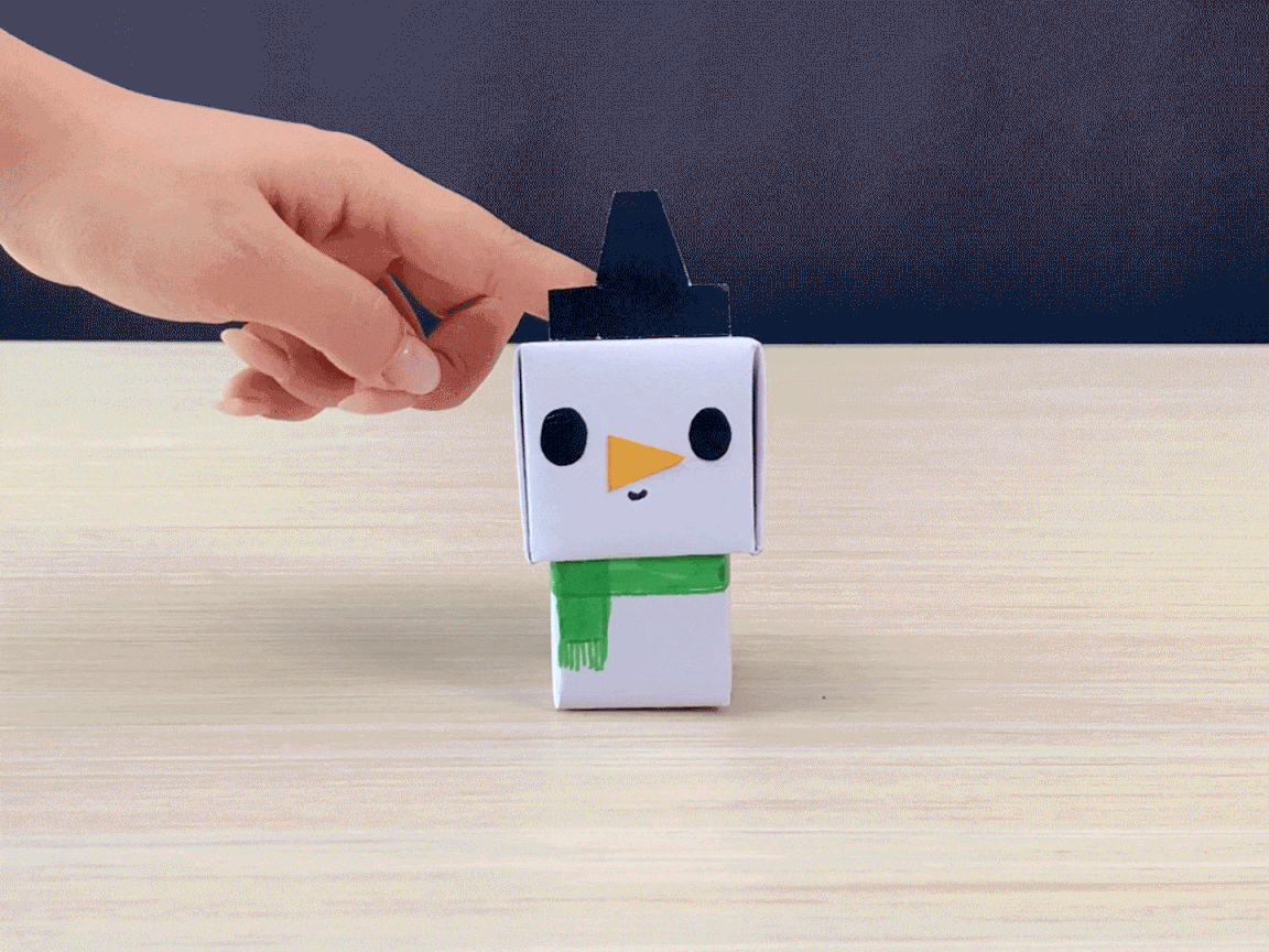 A small, cube-shaped paper snowman sits on a light wood table. It has black dot eyes, an orange triangle nose, and a light green scarf. A black, triangular hat rests on top. A hand gently taps the snowman's hat.