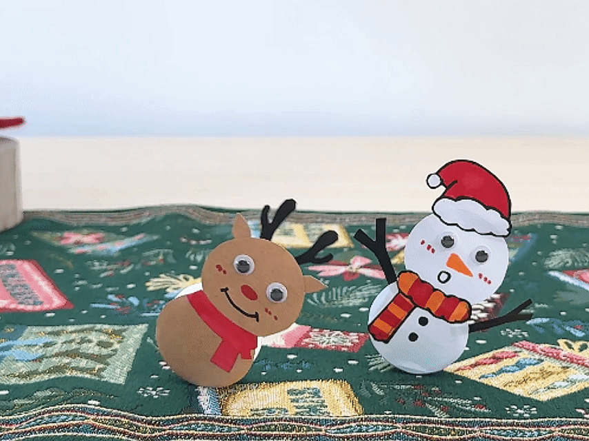Two charming Christmas figures, a reindeer and a snowman, wobble gently on a festive green tablecloth. The reindeer, crafted from brown paper, has black antlers, googly eyes, a cheerful red nose and mouth, and a red scarf. Next to it, the snowman, made of white paper, features googly eyes, rosy cheeks, an orange carrot nose, an orange and red striped scarf, black stick arms, and a classic red Santa hat. Both figures have rounded bases, causing their endearing wobble. A partially visible gift box sits in the background, enhancing the holiday scene.