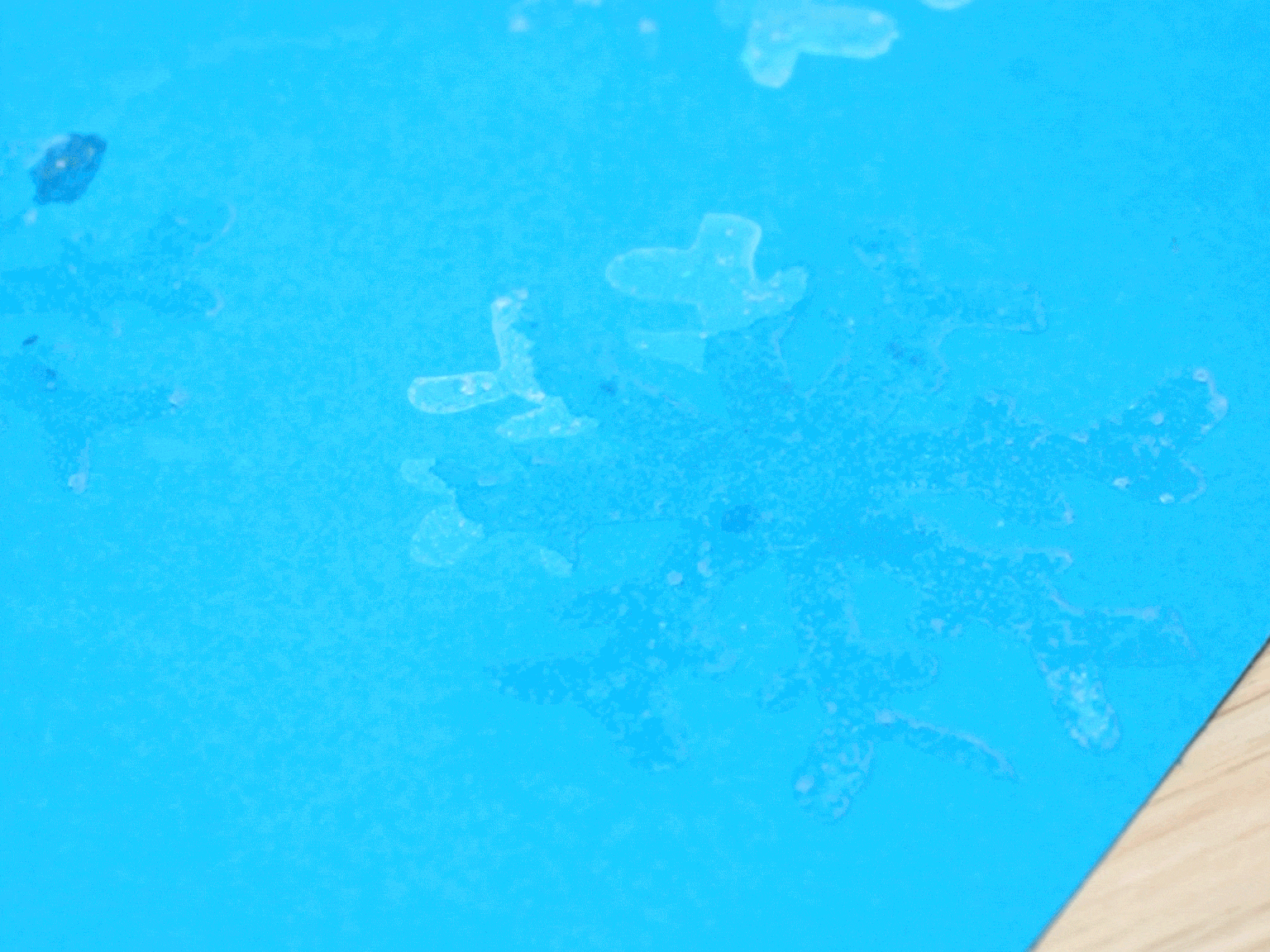 Several clear, glistening snowflake shapes are displayed on a vibrant blue piece of paper. The snowflakes appear to be made of a translucent, textured material, possibly glue or melted plastic, giving them a slightly raised and dimensional appearance. The light reflects off the snowflakes, creating a sparkling effect. The blue paper rests on a light wooden surface.