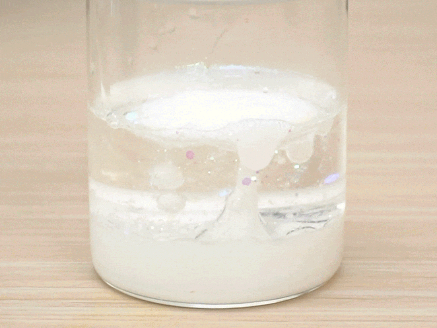 A clear glass jar, partially filled with a white substance, likely a powder or paste, sits on a light wooden table. A clear liquid, possibly water, is slowly added to the jar. As the liquid mixes with the white substance, it creates a cloudy, effervescent reaction. Small, iridescent particles, possibly glitter, are visible within the mixture, adding a subtle sparkle. The mixture gradually becomes more homogenous, with the white substance dissolving into the liquid.