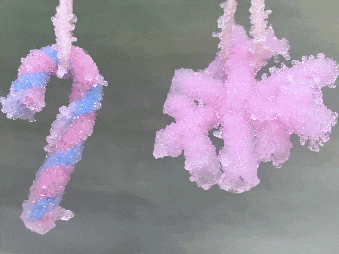 Two hanging ornaments, a candy cane and a snowflake, sparkle with a coating of pink crystals. The candy cane features pink and light blue stripes beneath the crystals. Both ornaments hang from thin twine against a muted gray-green background. The crystals catch the light, giving them a beautiful, glistening appearance.