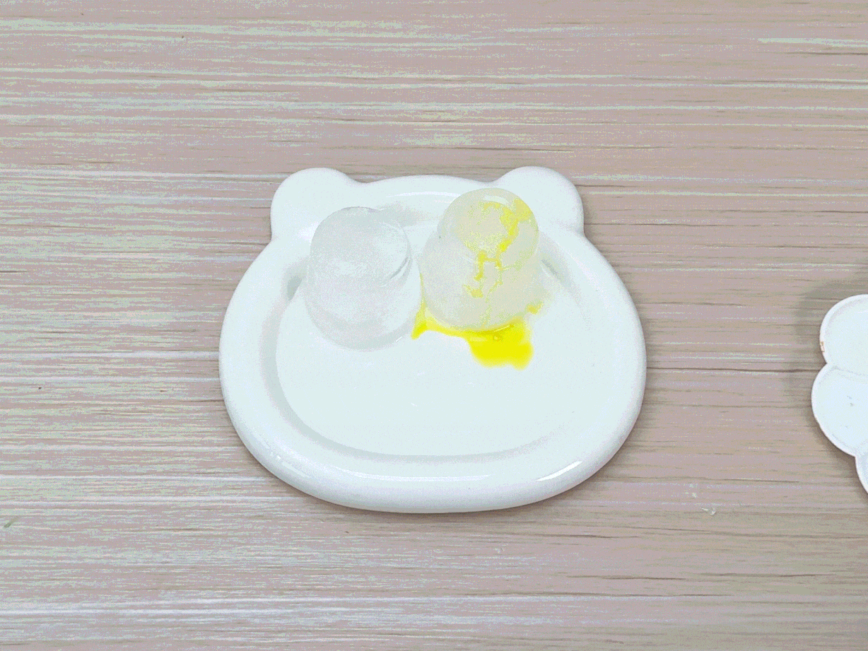 Two pieces of ice sit on a small, white, bear-shaped dish on a light wood table. One piece of ice is clear, while the other has thin, orange strands, possibly food coloring or yarn, embedded within it. A hand holding a paintbrush with green paint applies color to the clear ice. A small puddle of yellow liquid has collected around the base of the orange-streaked ice. A white paint palette is partially visible in the bottom right corner.
