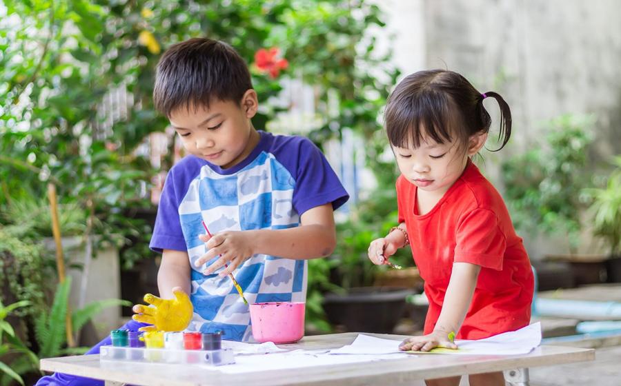 Two young children, a boy in a blue and white checked shirt and a girl in a red dress, are engrossed in a painting activity outdoors, surrounded by lush greenery, happily creating artwork with paint and their hands.