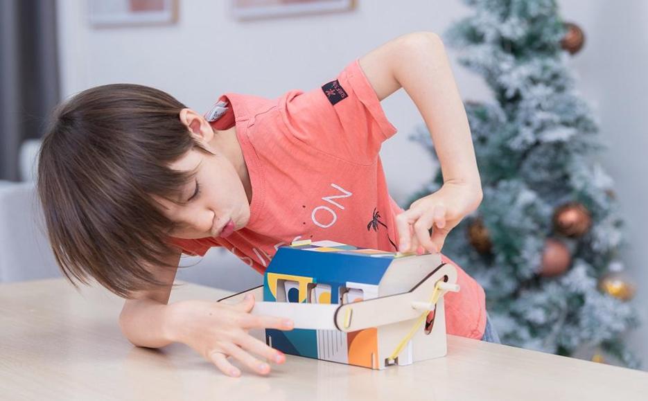 A young boy, engrossed in assembling a colourful, small-scale model house with moving parts, leans over the table, carefully manipulating the pieces with focused concentration.