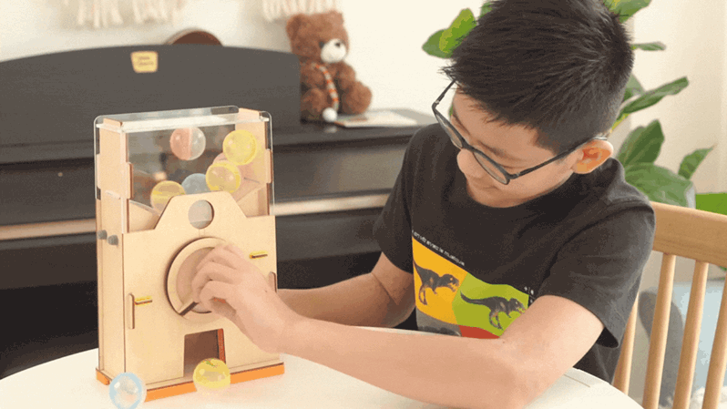 A boy wearing glasses operates a handcrafted wooden ball dispenser, turning a crank to release colourful balls from a clear acrylic container; a fun and engaging hands-on activity.