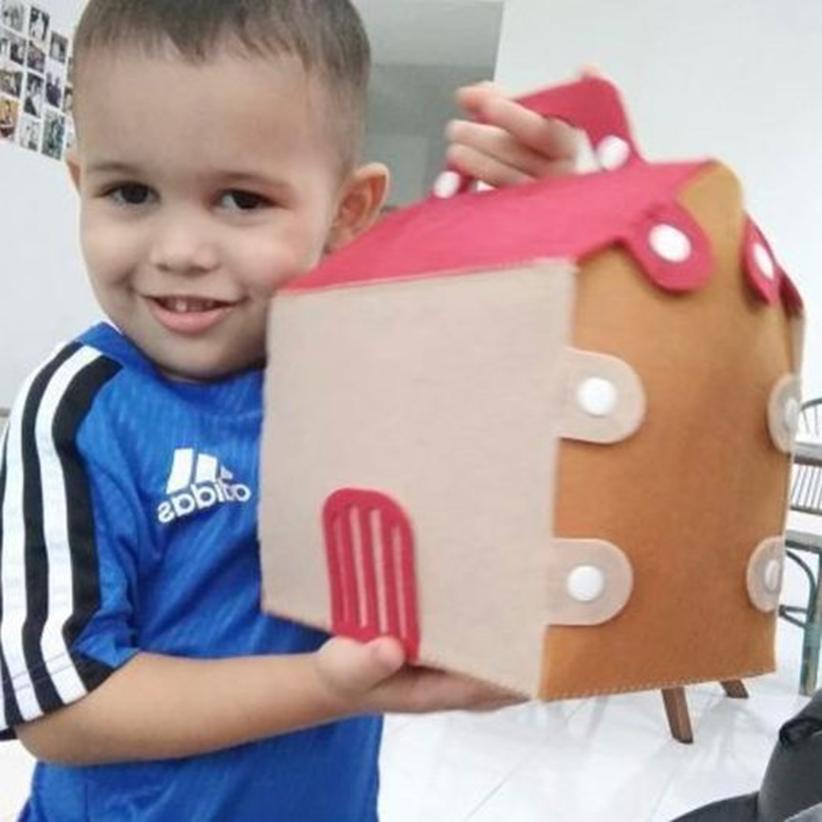 A young boy proudly displays a handcrafted felt house, featuring a red roof and small door, showcasing a completed creative project.