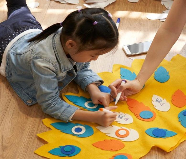 A young girl lies on the floor, meticulously decorating felt peacock feathers affixed to a larger yellow felt piece with white markers, assisted by an adult hand also using a marker, creating a collaborative craft project.