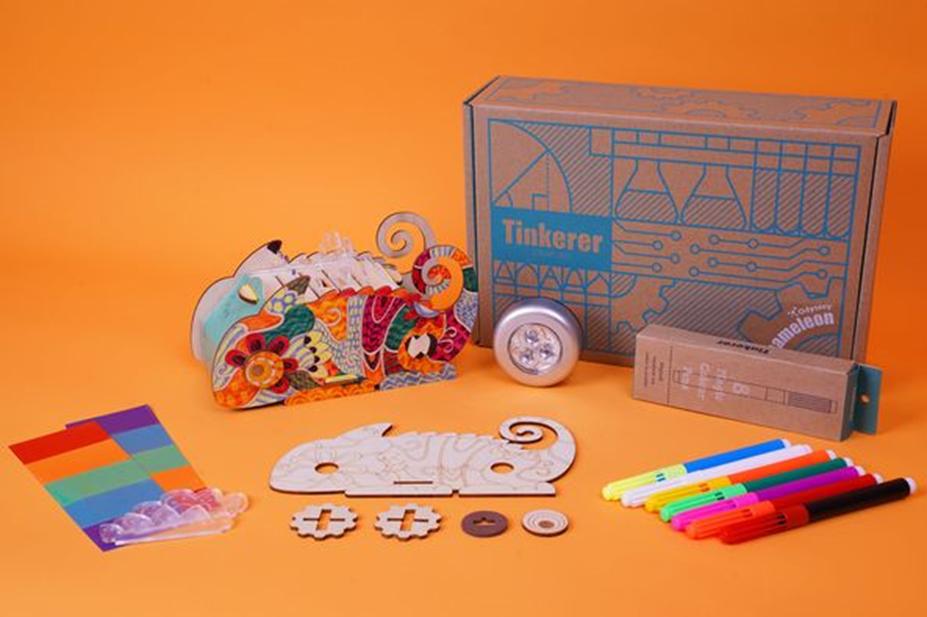 A colourful chameleon-themed craft kit is laid out, showcasing its contents: a partially assembled chameleon model, coloured markers, a small LED light, and various other components, all ready for a creative building and decorating activity.